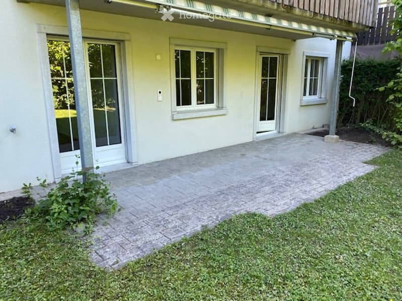 A paved patio area adjacent to a house with white walls and large windows, surrounded by a small garden with grass and some plants.