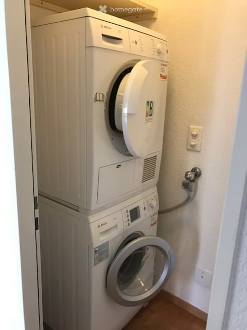 Compact bathroom with stacked Bosch washing machine and dryer unit, white tiled walls, wooden floor, and light switch.