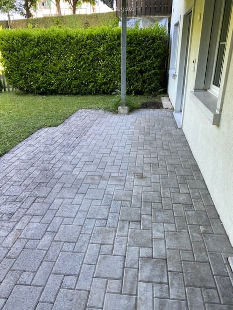 Brick patio area adjacent to a house, with neatly trimmed hedges and a grassy lawn in the background.