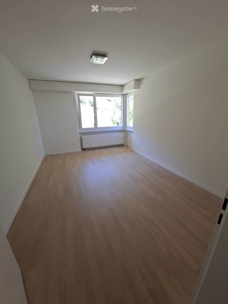 Empty room with wooden flooring, white walls, and large windows with natural light. Features a radiator and a ceiling light fixture.