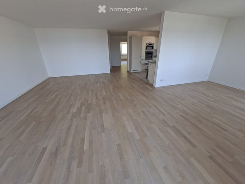 Empty room with wooden flooring and white walls, featuring a small kitchen area with cabinets and appliances visible in the background.