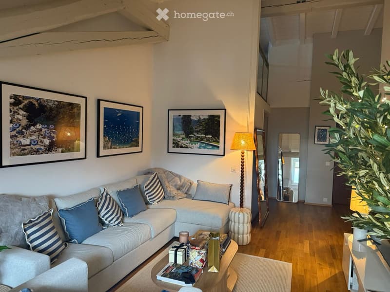 Cozy living room with a light grey sectional sofa, blue and white striped cushions, a wooden coffee table with books and gifts, a floor lamp, and a large plant in the corner.