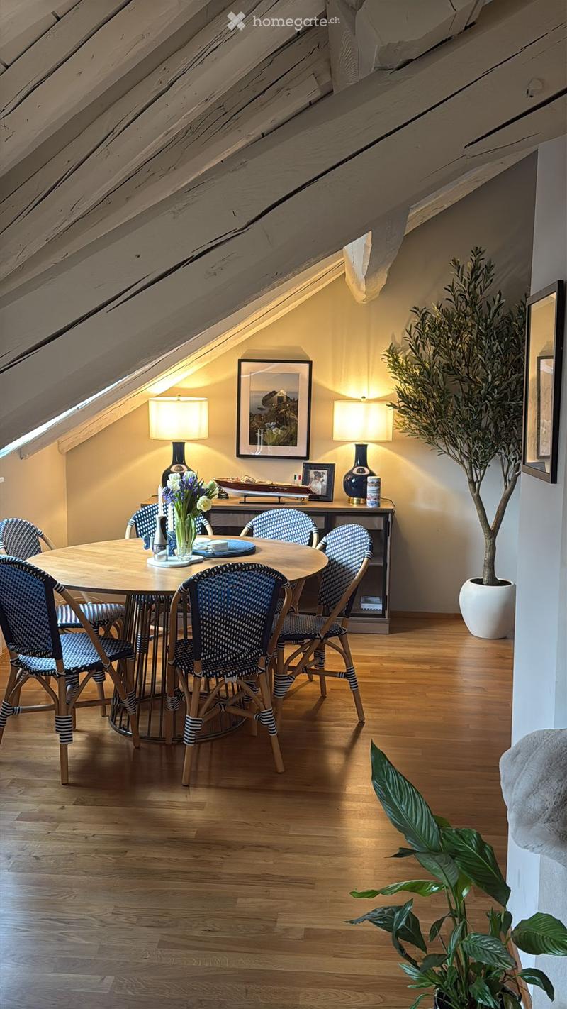 Cozy attic dining area with wooden floor, round wooden table surrounded by six striped chairs, two table lamps, decorative picture frames, and potted plants.