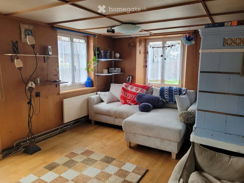 Cozy living room with wooden flooring, a sofa with colorful pillows, a small table with shelves, a floor lamp, and a plant near the window.