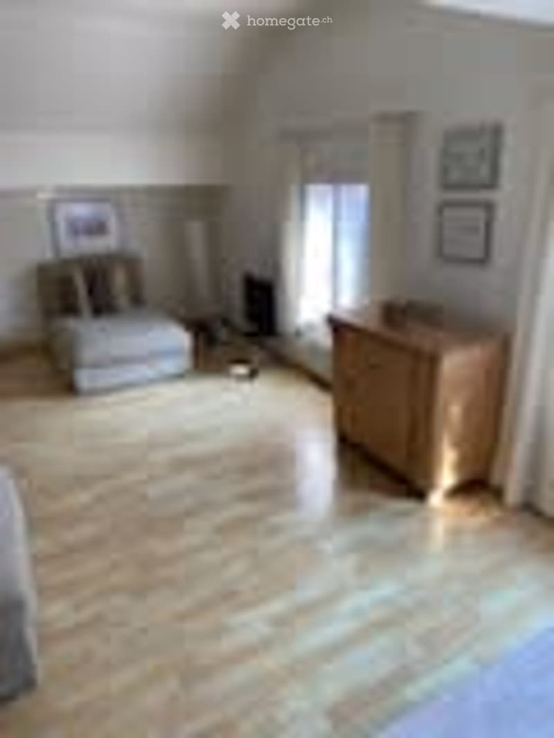 Bright living room with light wooden flooring, white walls, large window with blinds, a white sofa, a wooden chair, and a small table with picture frames on the wall.