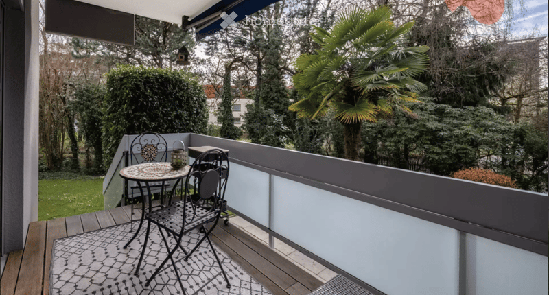 Balcony with wooden decking, modern railing, small round table with two chairs, decorative lantern, and lush greenery in the background.