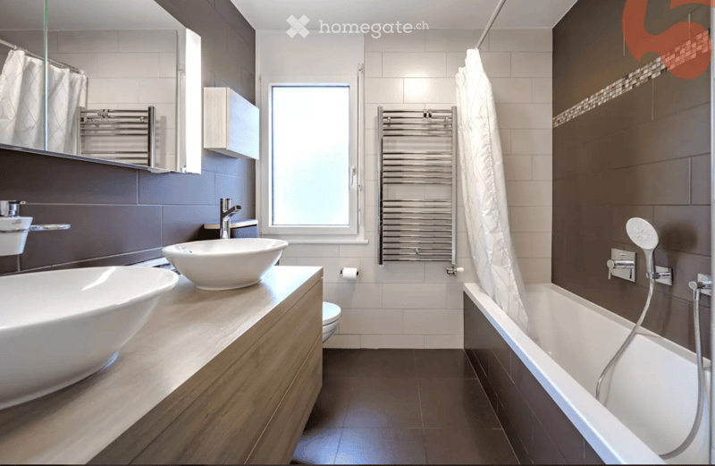 Modern bathroom with a large bathtub, separate sink, and a shower area. Features include a dark tiled wall, a wooden vanity, a towel, and a radiator.