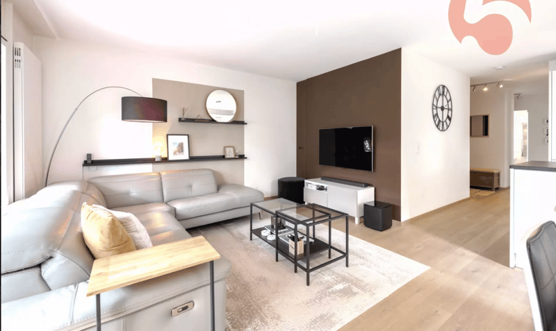 Modern living room with a grey sectional sofa, wooden coffee table, floor lamp, wall-mounted TV, and decorative shelves.