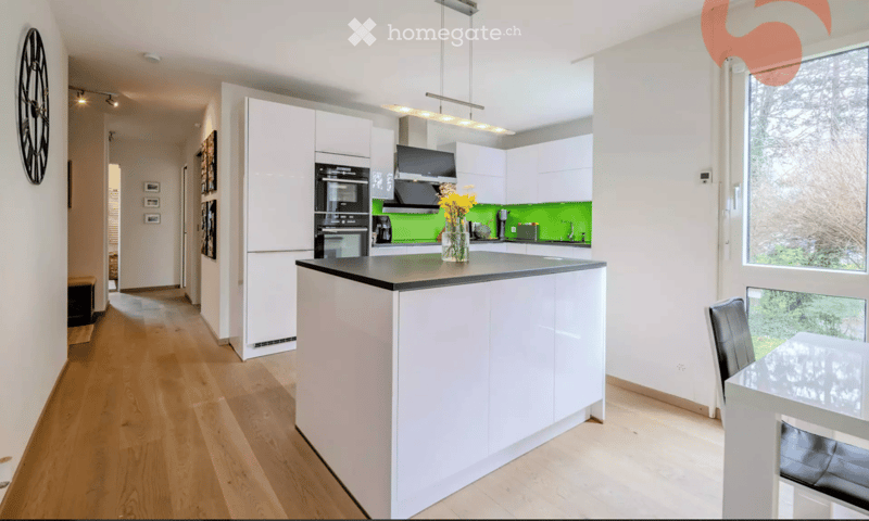 Modern kitchen with white cabinets, a central island, stainless steel appliances, and a green accent wall. Features a dining area with a glass table and black chairs.
