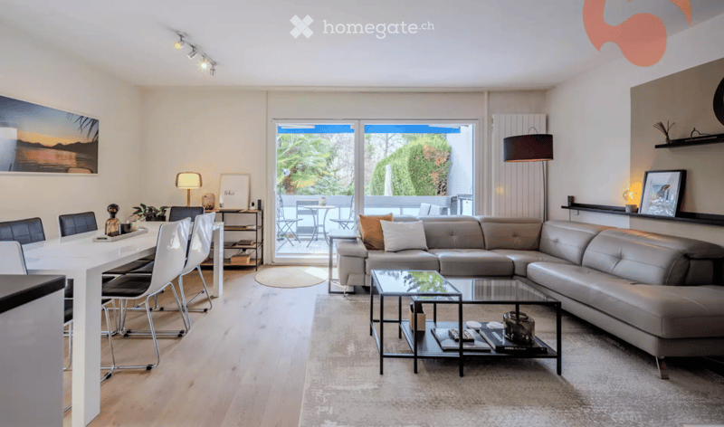 Modern living and dining area with a grey sectional sofa, glass coffee table, white dining table with chairs, and large windows opening to a patio.