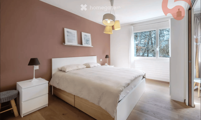 Modern bedroom with a large bed, nightstand, stool, and wall-mounted picture frames. The room features wooden flooring and large windows with a view of trees.
