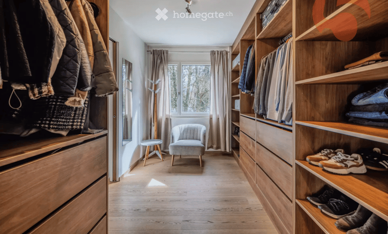 A well-organized walk-in closet with wooden shelves and drawers, a small seating area with a grey armchair and a round stool, and a coat rack near the window.