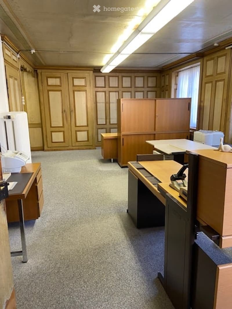 Office space with multiple desks, chairs, cabinets, and a printer. The room has wooden paneling and large windows with blinds.