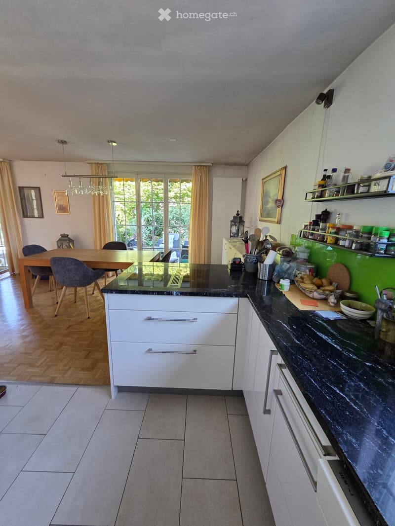 Modern kitchen with white cabinets, black countertops, and a dining area with a wooden table and chairs. Shelves hold various kitchen items and decorations.