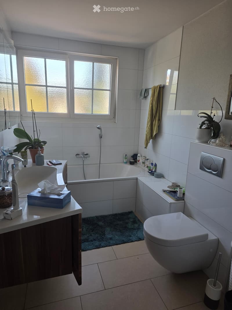 Modern bathroom with white tiles, a bathtub, a sink with a faucet, a toilet, a towel hanging on the wall, and a small plant on the windowsill.