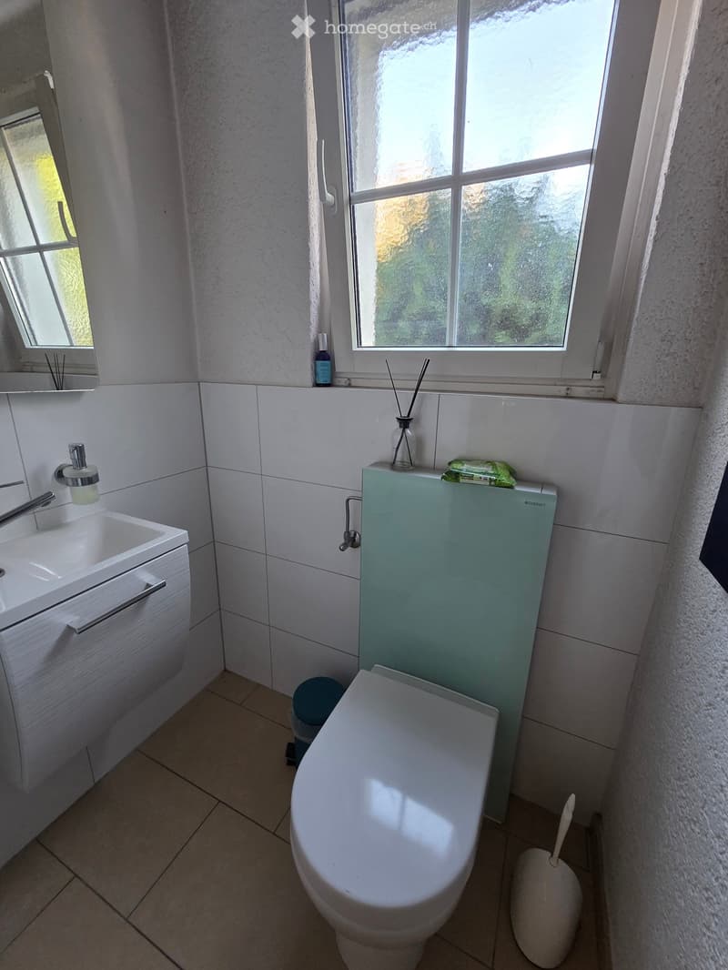 Modern bathroom with white tiled walls, a toilet, a sink with a mirror, and a small window letting in natural light.