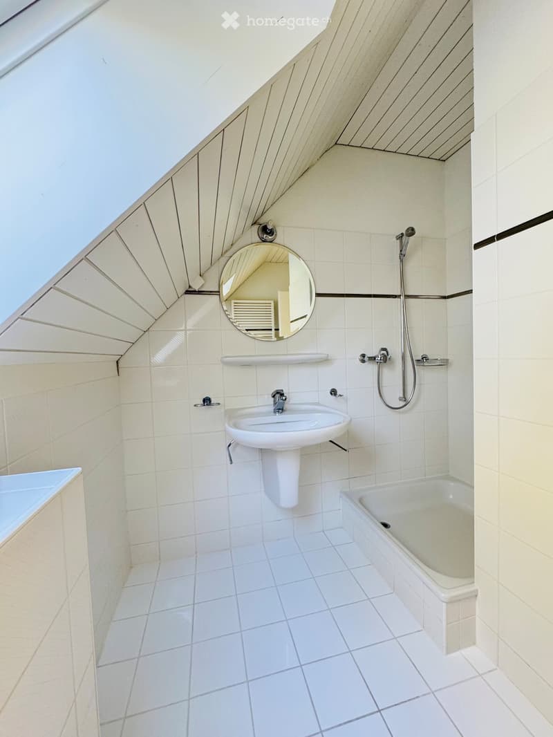 Modern bathroom with white tiles, a sink, bathtub, mirror, and showerhead. The ceiling has a skylight and wooden paneling.