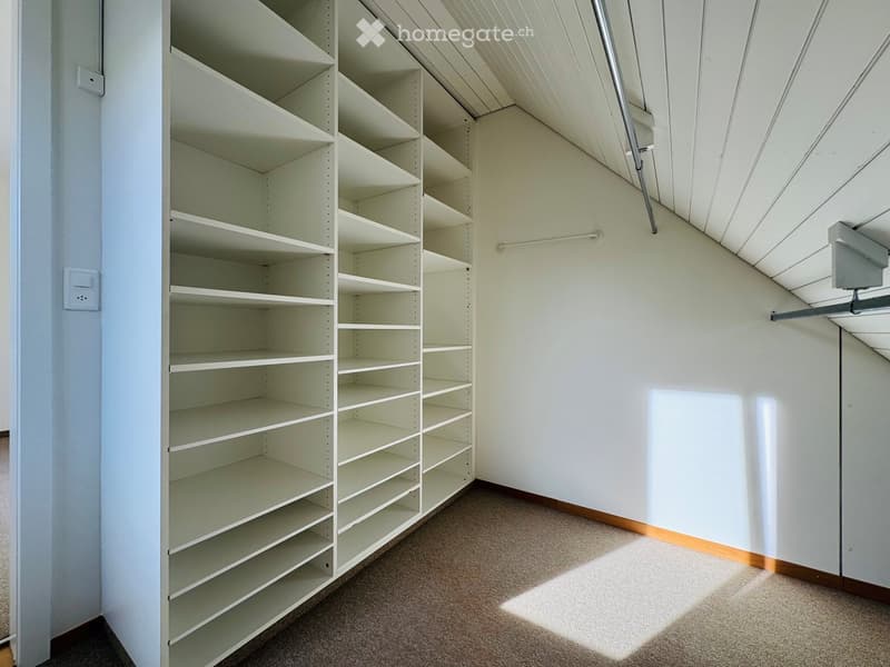 Empty attic space with built-in white shelving units and a slanted ceiling. Sunlight streams in from a window on the right side.