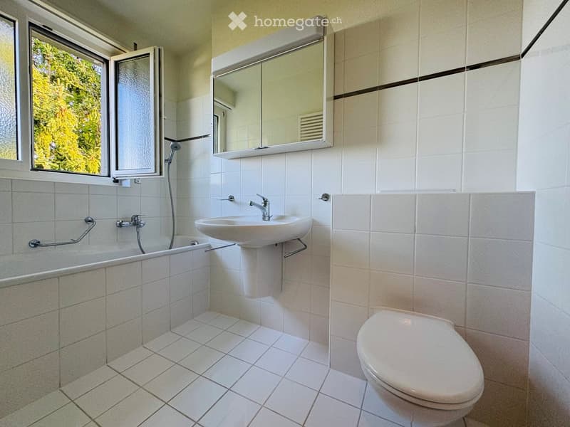 Modern bathroom with white tiles, a bathtub, a sink with a faucet, a toilet, and a large mirror above the sink. The room has a window with a view of greenery.