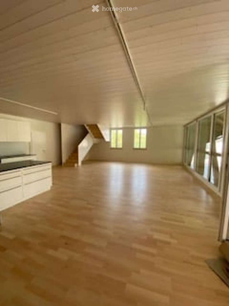 Bright, open-plan room with wooden flooring, white walls, and large windows. The space is completely empty, with no furniture or equipment visible.