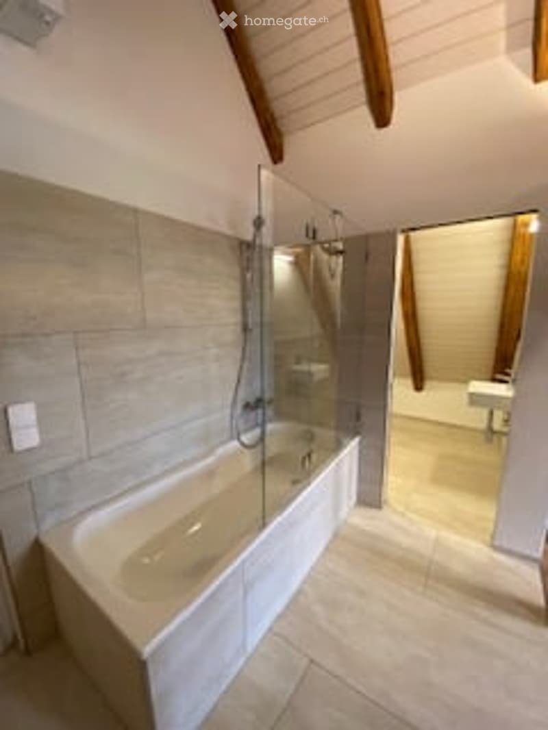 Modern bathroom with a large bathtub, glass shower enclosure, and a large mirror. The walls are tiled in a light grey color, and the ceiling has exposed wooden beams.