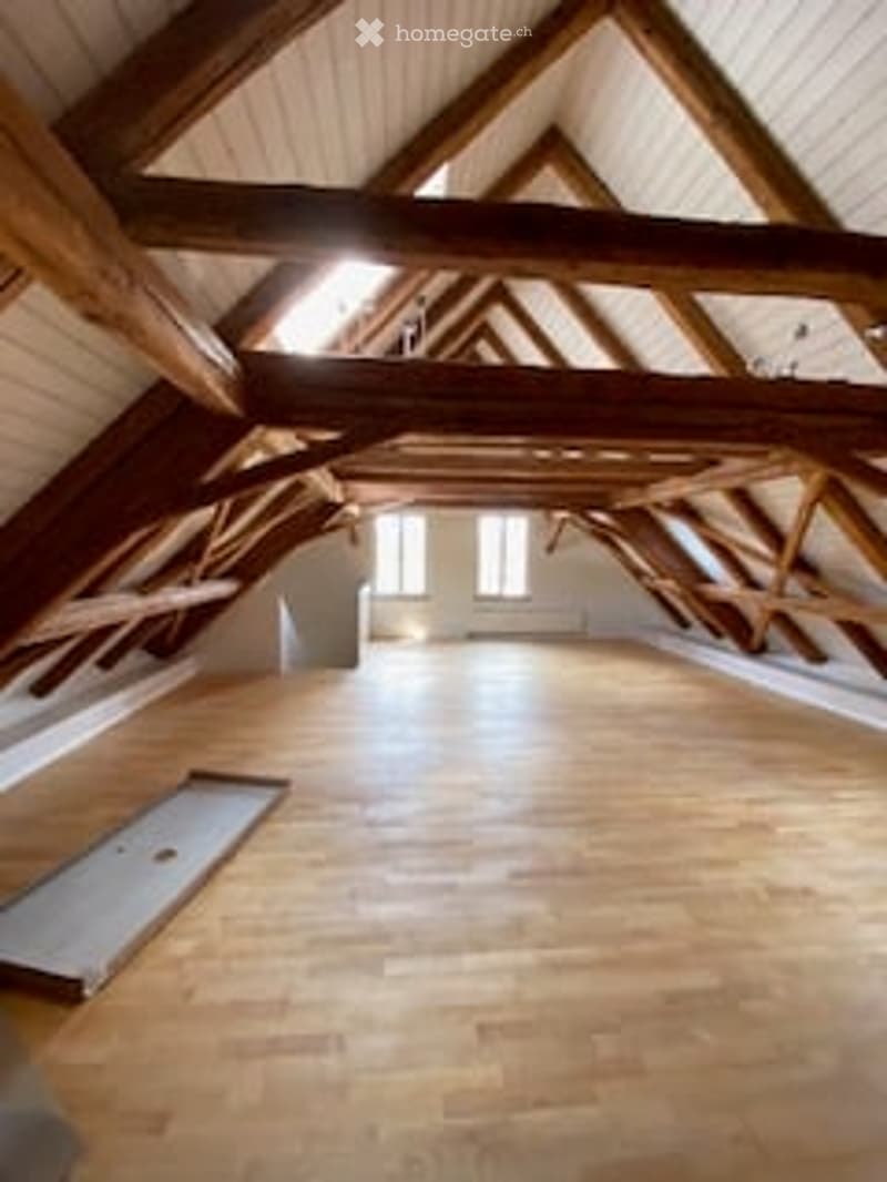 An attic space with exposed wooden beams and a wooden floor, featuring large windows allowing natural light to enter.