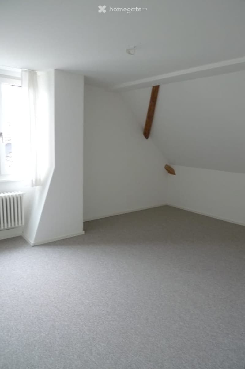 Empty room with white walls, grey carpet, and a window with white curtains. No furniture or equipment present.