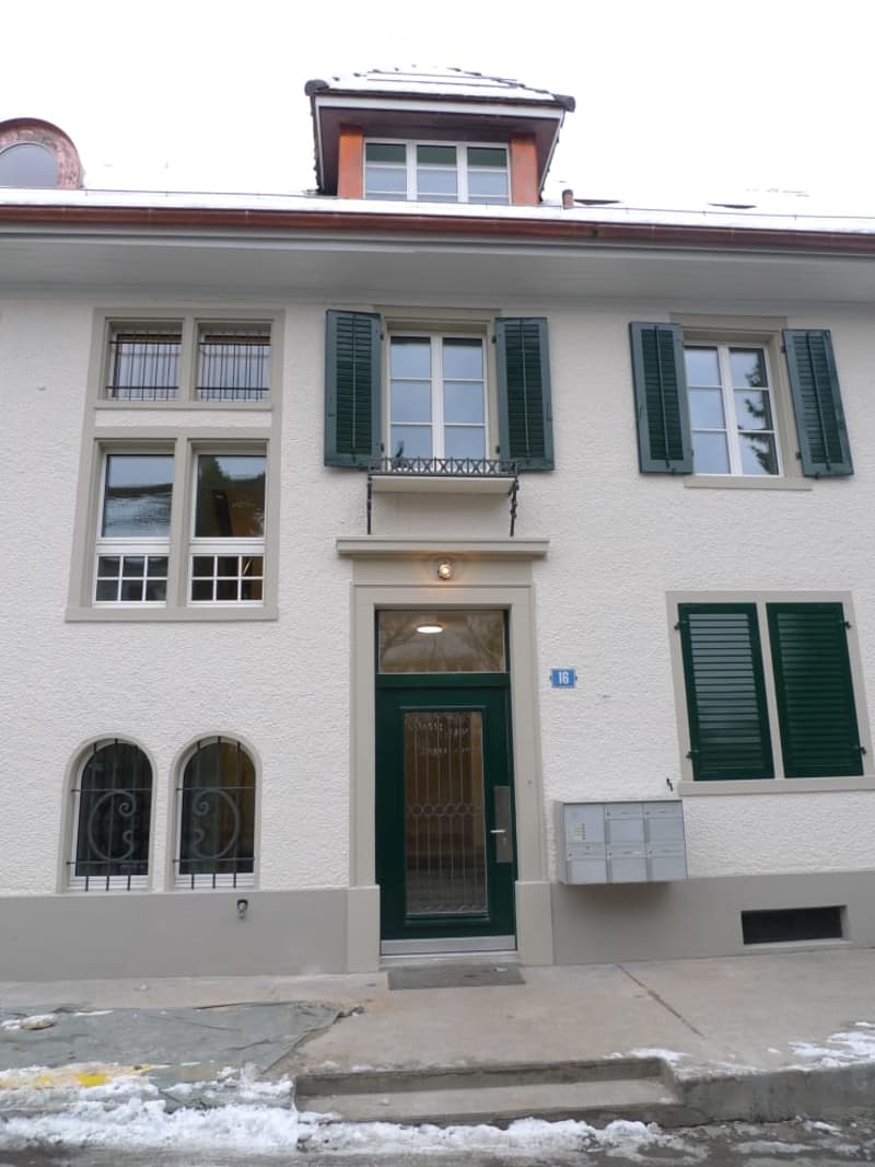 A two-story residential building with white walls, green shutters, and a green front door. The entrance features a small balcony above the door, and the building has multiple windows with metal bars on some.