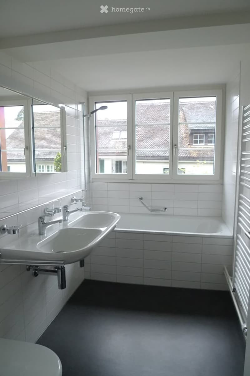 Modern bathroom with white tiles, double sink, bathtub, large window, and mirror.