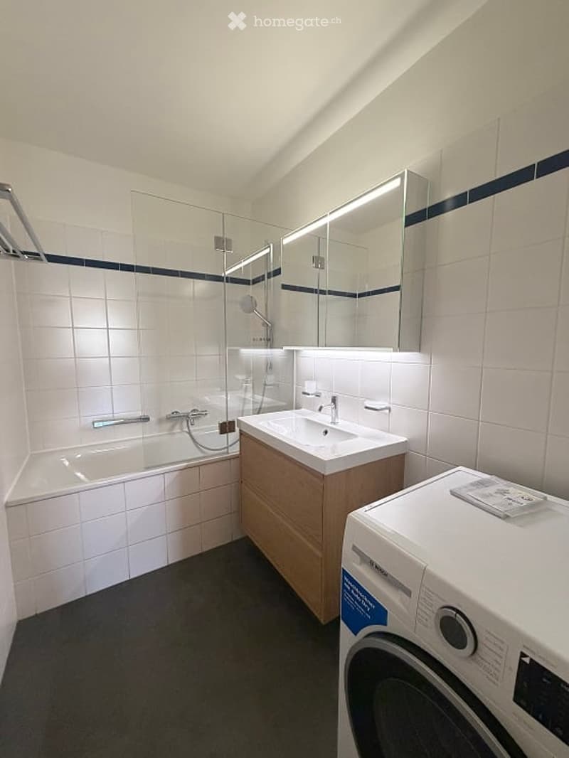 Modern bathroom with white tiled walls, a bathtub, a sink with a wooden cabinet, a large mirror, and a washing machine in the corner.