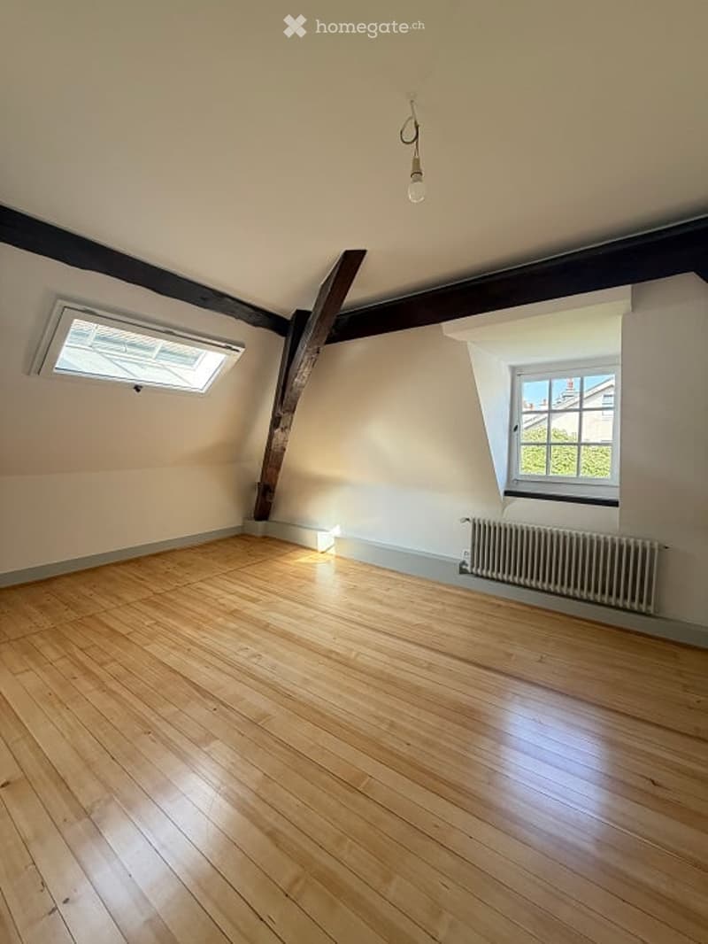 Empty attic room with wooden flooring, exposed wooden beams, and two windows allowing natural light.