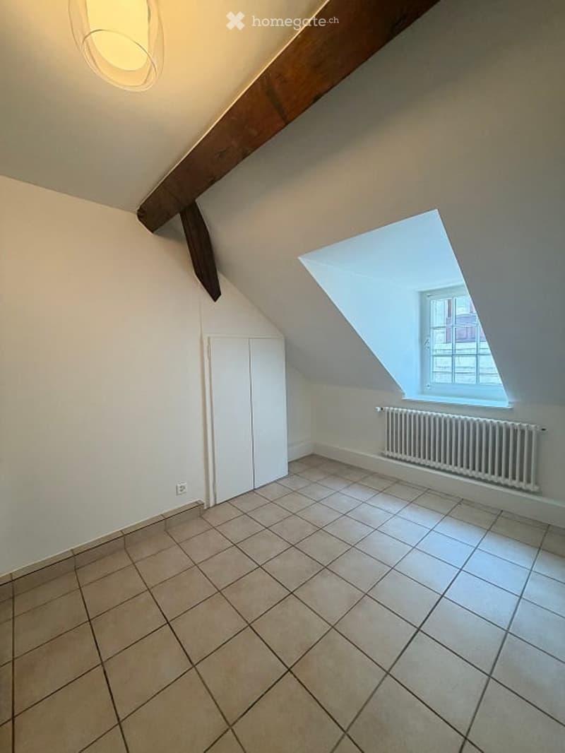 Empty attic room with exposed wooden beams, tiled floor, white walls, built-in closet, and a skylight window.