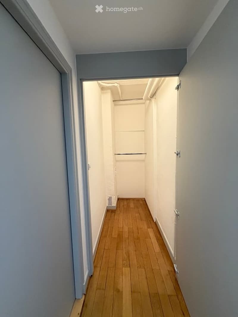 A narrow, empty hallway with light wooden flooring and white walls. The hallway features a closet with a single rod and a small vent on the wall.