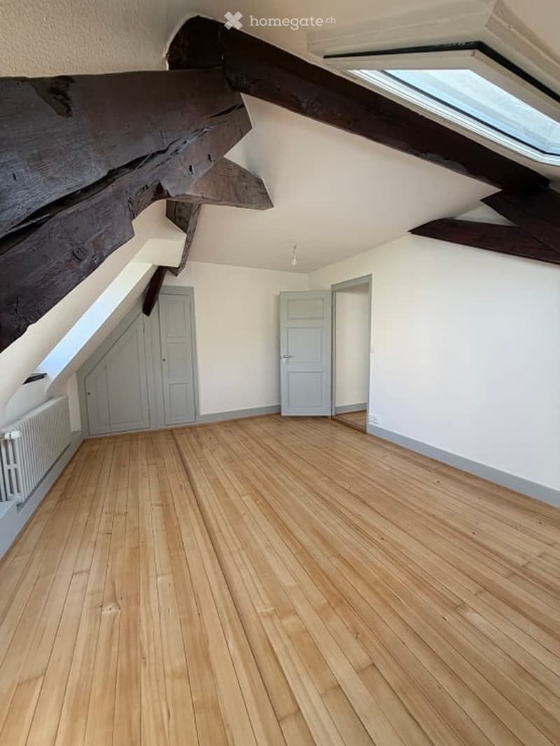 Empty attic room with wooden flooring, white walls, exposed wooden beams, and a skylight window. Two white doors are visible.