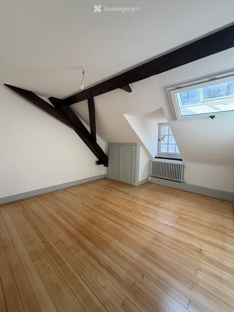 Empty attic room with wooden floor, exposed wooden beams, white walls, and a small built-in closet.