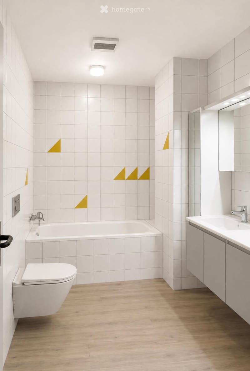 Modern bathroom with white tiled walls, wooden floor, bathtub, toilet, sink, and mirror. Features geometric yellow wall accents and a glass shower enclosure.
