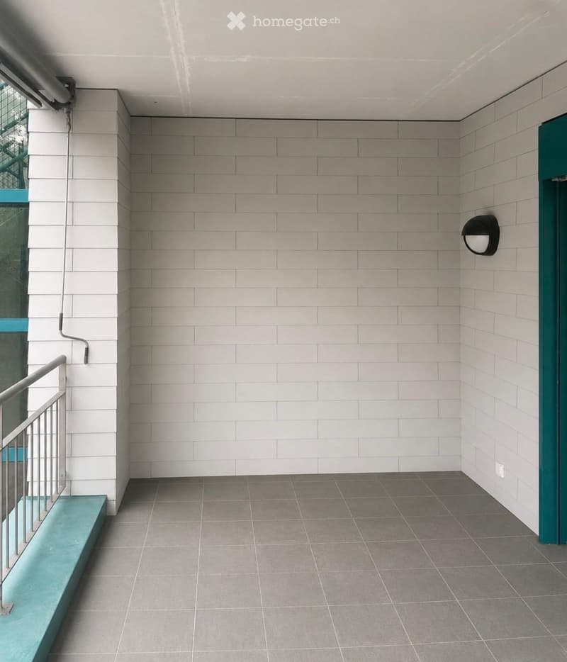 Modern hallway with grey tiled walls and floor, metal railing, teal-colored bench, and a wall-mounted light fixture.