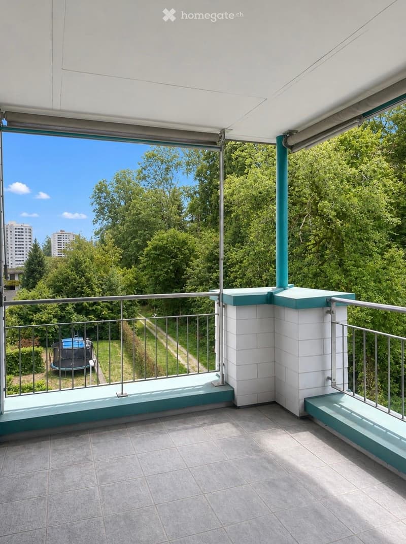 Balcony with tiled floor, metal railing, and retractable awning, overlooking a green area with trees and a pathway.