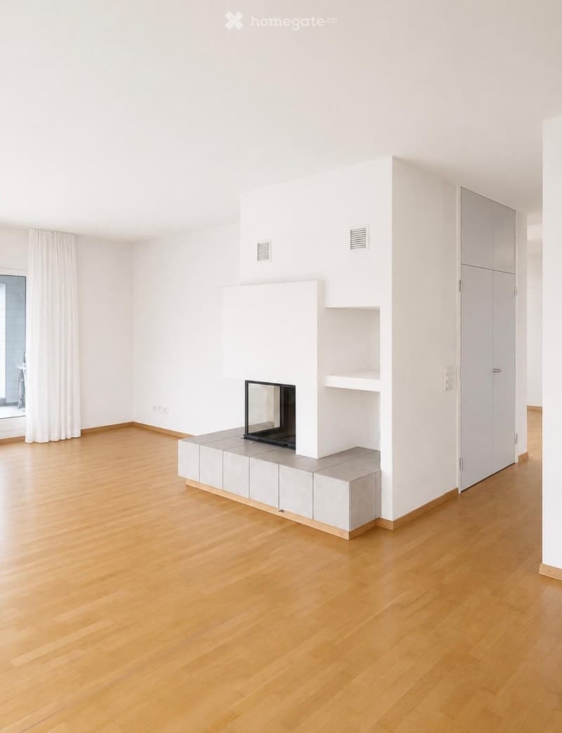 Empty room with light wooden flooring, white walls, and a built-in fireplace with a mirror above it. The room has large windows with white curtains and a door leading to another space.