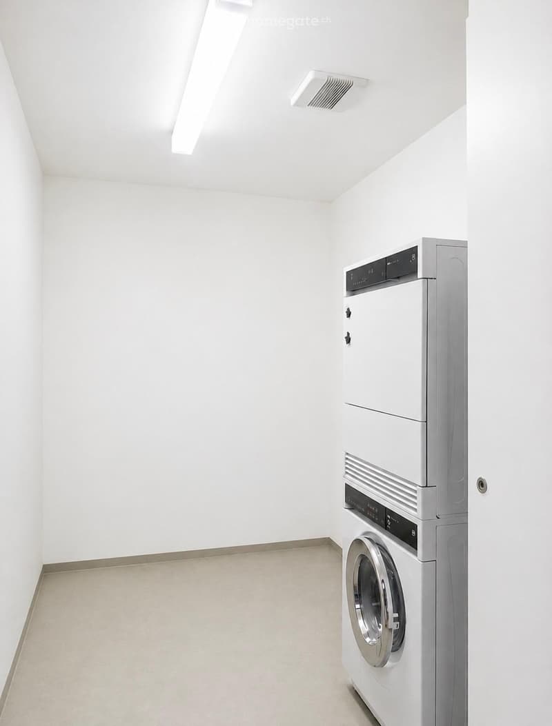 A small, clean room with white walls and a light-colored floor, featuring a stacked washing machine and dryer unit.