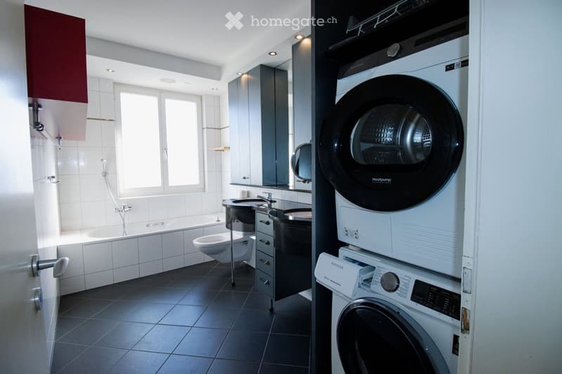 Modern bathroom with a bathtub, toilet, sink, mirror, and a stacked washing machine and dryer unit.