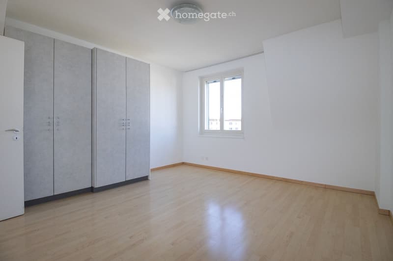 Empty room with light wooden flooring, white walls, and a large window letting in natural light. Features built-in grey wardrobes and a modern ceiling light fixture.