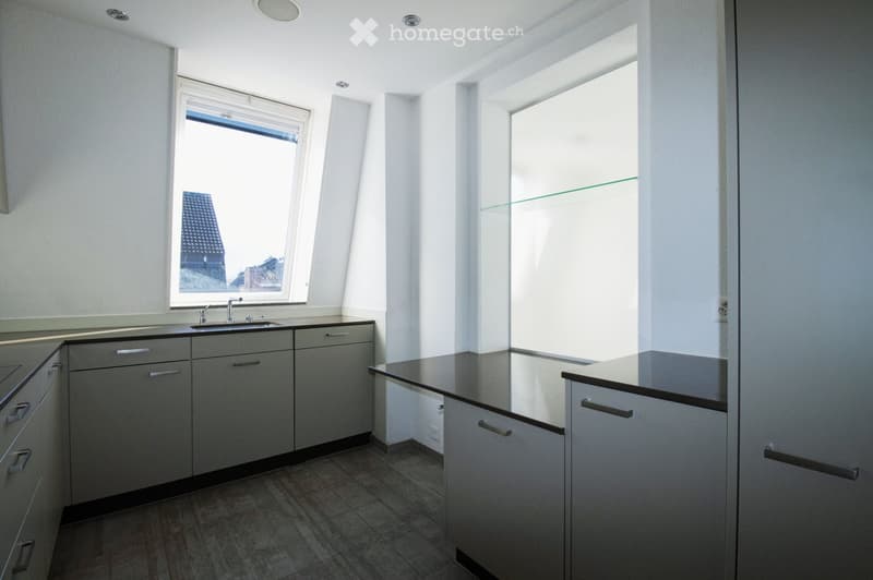 Modern kitchen with grey cabinets, a black countertop, and a sink under a window. The space is bright with natural light from multiple windows, including one with a view of a building exterior.