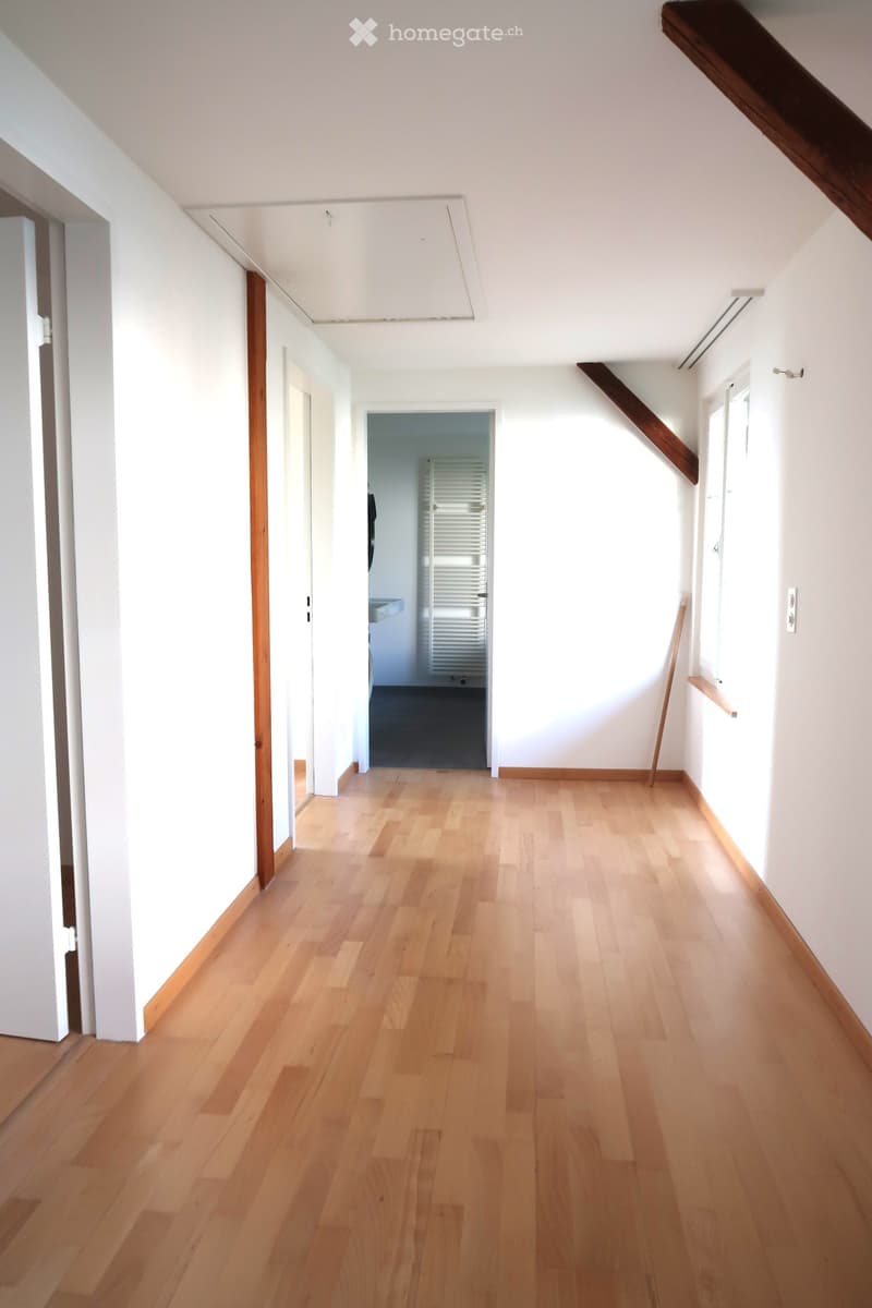 Bright, empty hallway with light wooden flooring, white walls, and a slanted ceiling. Features include a small window on the right and a doorway leading to another room.