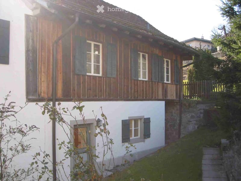 Two-story house with wooden facade and white lower section, featuring multiple windows with shutters and a small balcony on the upper level.