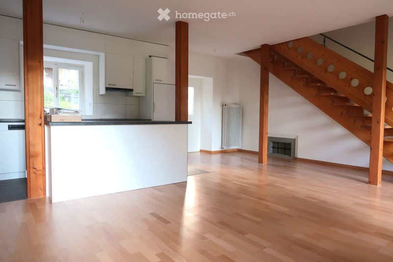 Open-plan living and kitchen area with light wooden flooring, white cabinets, a refrigerator, and a dishwasher. A wooden staircase leads to an upper level.