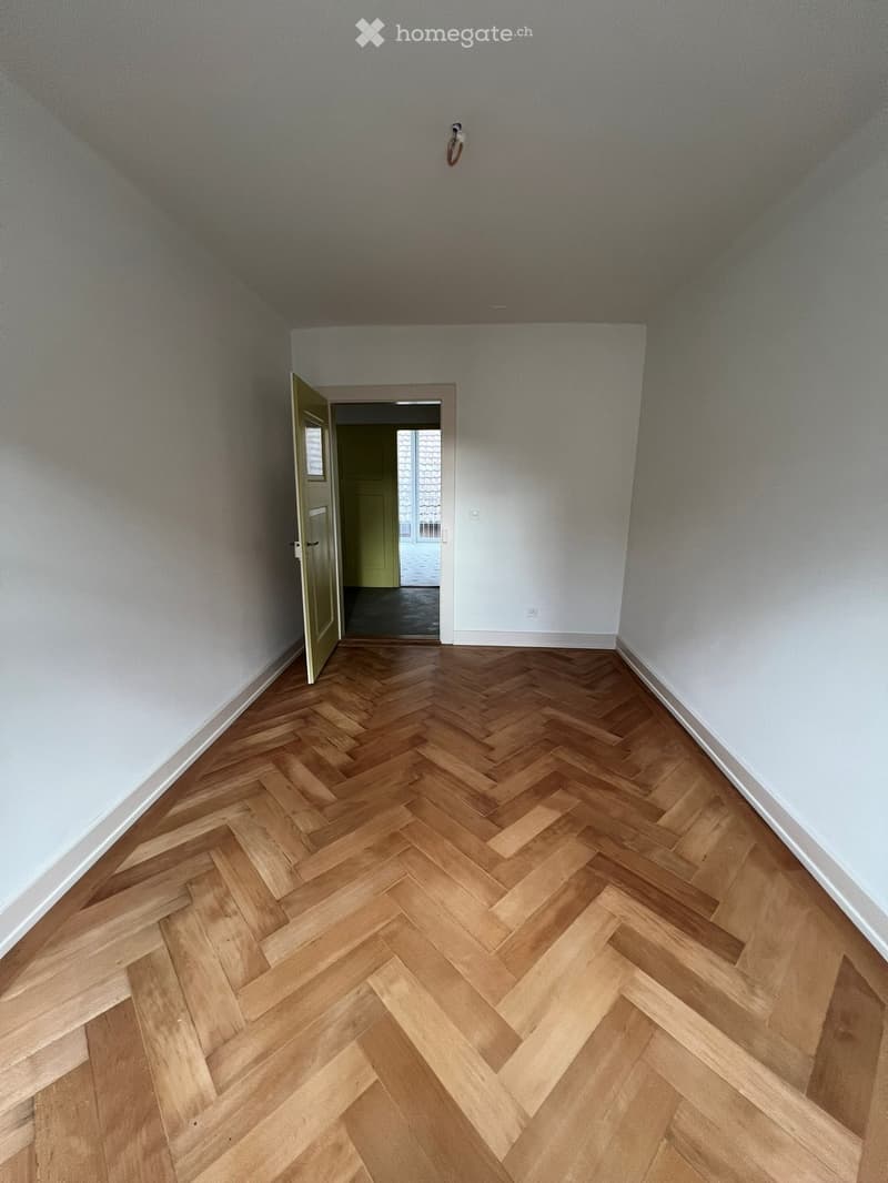 Empty hallway with herringbone wooden flooring, white walls, and an open door leading to another room.