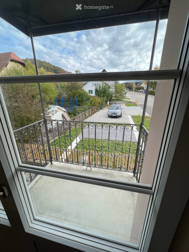 Balcony with metal railing overlooking a driveway, parked car, and surrounding greenery.