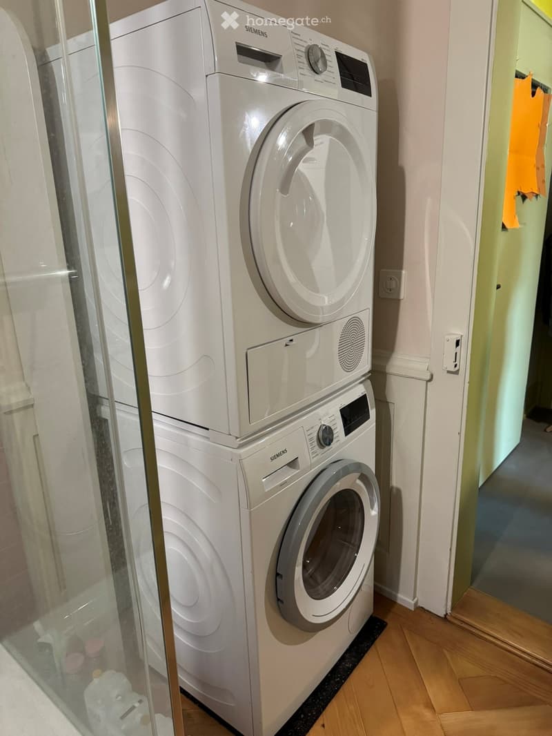 A small room with a white washing machine and dryer stacked vertically, placed on a wooden floor next to a glass door.