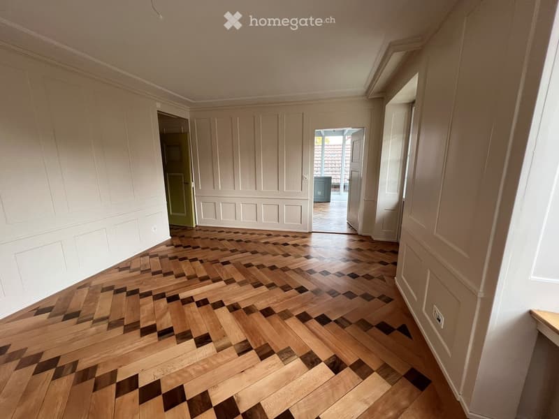 Empty hallway with wooden herringbone flooring and white paneled walls, featuring several doors and an open doorway leading to another room.